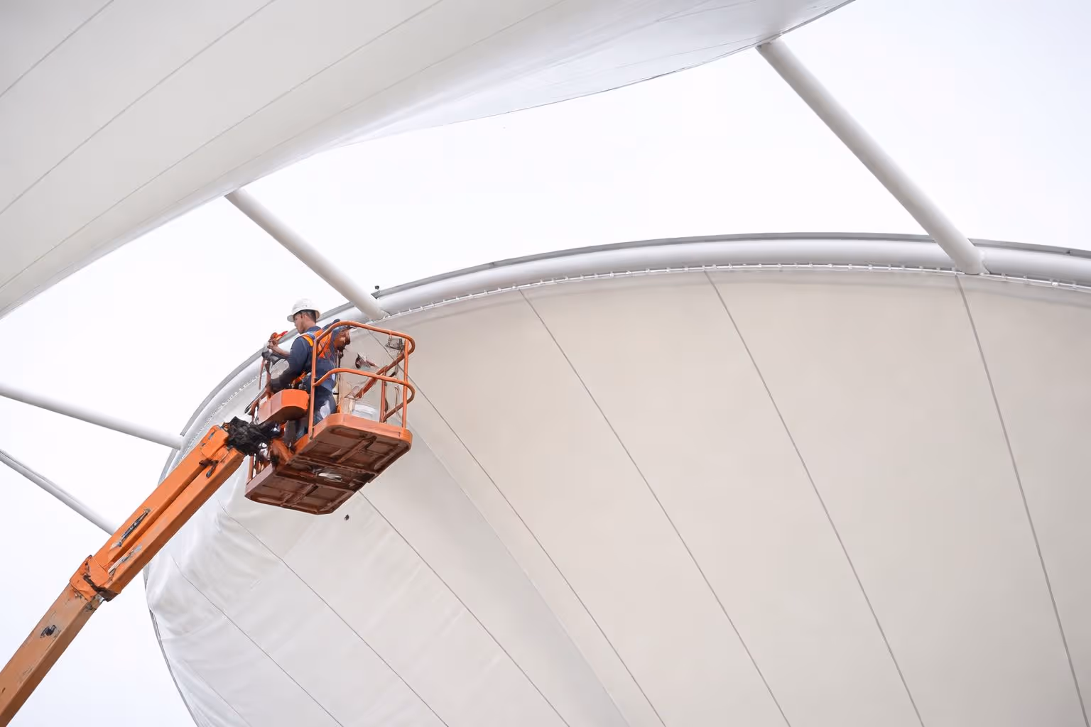 Two workers in safety gear on an orange aerial lift inspecting or repairing a large white tensile fabric structure.